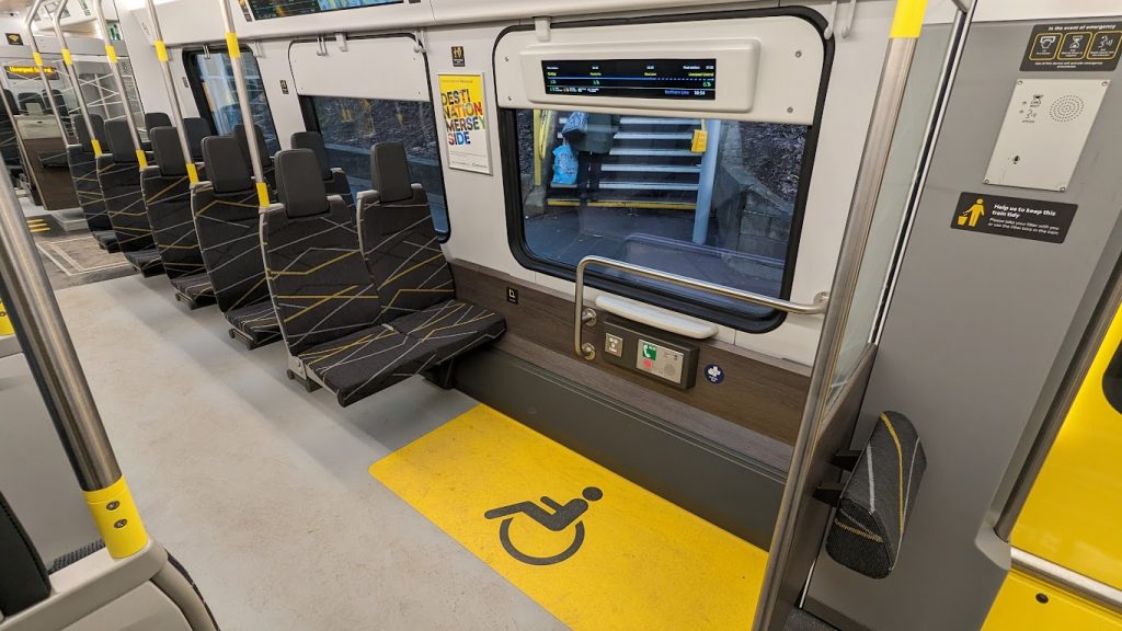 The wheelchair space on a class 777 train. It is marked on the floor with a yellow box and a black wheelchair symbol. Adjacent to the space is a handrail, plug socket (with USBs), a small table and a call for aid alarm.